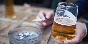 Foto: Eine Hand h&auml;lt ein halb gef&uuml;lltes Bierglas und h&auml;lt eine Zigarette, daneben steht ein Aschenbecher.