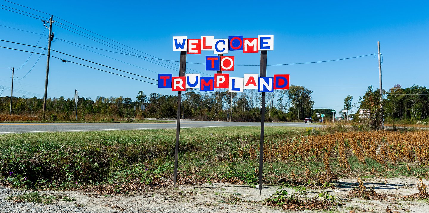 Foto: "Welcome to Trumpland" flattert in Großbuchstabenfähnchen in den Farben Weiß/Blau/Rot im Wind vor einem Feld, daneben eine Straße, auf der ein Auto fährt.