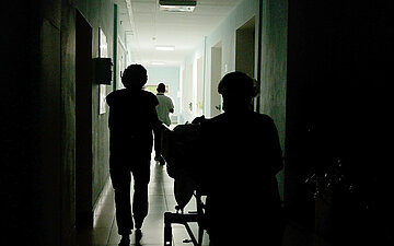 Dunkler Krankenhausflur mit mehreren Personen als Silhouetten. Zwei Personen schieben ein Krankenbett in Richtung eines hell erleuchteten Bereichs.