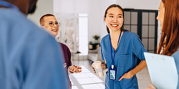 Mehrere Pflegekr&auml;fte in blauer und violetter Arbeitskleidung unterhalten sich l&auml;chelnd in einem hellen Krankenhausbereich. Eine Pflegekraft h&auml;lt einen Kaffeebecher, im Vordergrund ist ein ge&ouml;ffneter Laptop zu sehen.