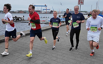 Verschiedene L&auml;uferinnen und L&auml;ufer, jeweils mit ihren Startnummern an den T-Shirts, laufen vor der maritimen Kulisse der Kiellinie den letzten Streckenabschnitt bis zum Ziel. 