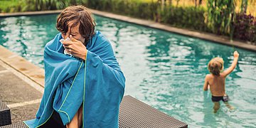Leisure Sickness Ein Mann sitzt mit einer Decke an einem Pool und putzt sich die Nase, während im Hintergrund ein Kind im Pool spielt.