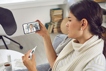 Eine Frau sitzt an einem Tisch, dort steht eine Tasse mit einer Fl&uuml;ssigkeit, daneben liegt ein Medikament. In der linken Hand h&auml;lt sie ein Fieberthermometer, in der rechten h&auml;lt sie ihr Smartphone, auf dem im Display eine &Auml;rztin sichtbar ist, mit der sie telefoniert. 