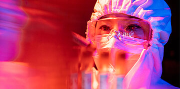 Eine Person in Schutzkleidung und Schutzbrille arbeitet konzentriert im Labor mit Proben und Ger&auml;ten unter farbigem Licht.