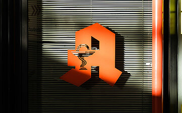 Rotes Apotheken- &bdquo;A&ldquo; mit Schale-und-Schlange-Symbol, teilweise verschattet, vor einem dunklen Hintergrund.