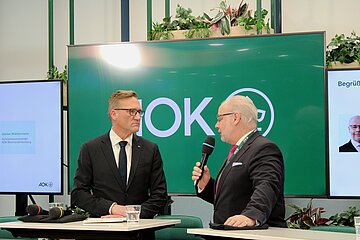 zwei M&auml;nner stehen vor einem gr&uuml;nen Hintergrund, einer h&auml;lt ein Mikrofon