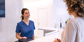 Eine Frau in OP-Kleidung steht hinter einer Empfangstheke um Patienten zu begr&uuml;&szlig;en.