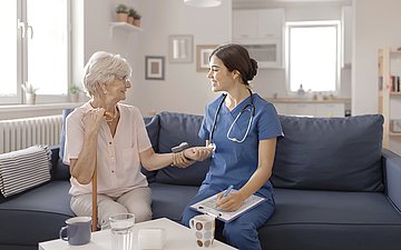 Pflegebegutachtung Eine Gutachterin sitzt neben einer alten Frau zu Hause auf dem Sofa, auf ihrem Schoß einen Fragebogen.