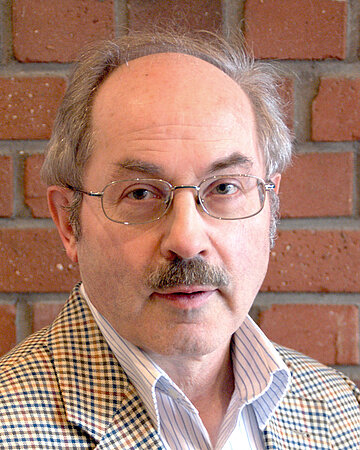 Professor Hirsch ist ein Mann in mittleren Jahren. Er steht vor einer Backsteinmauer. Er tr&auml;gt eine Brille und ein Oberlippenbart. Sein Jackett ist kariert und er tr&auml;gt darunter ein helles Hemd.