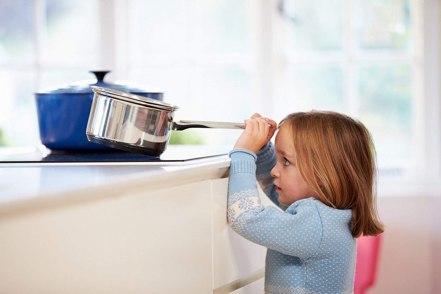 Das Foto zeigt ein kleines Kind, das vor einem Kochfeld steht, gerade so über die Kante schauen kann schauen und mit beiden Händen nach einem Topf mit Stilgriff greift, der auf dem Kochfeld steht.