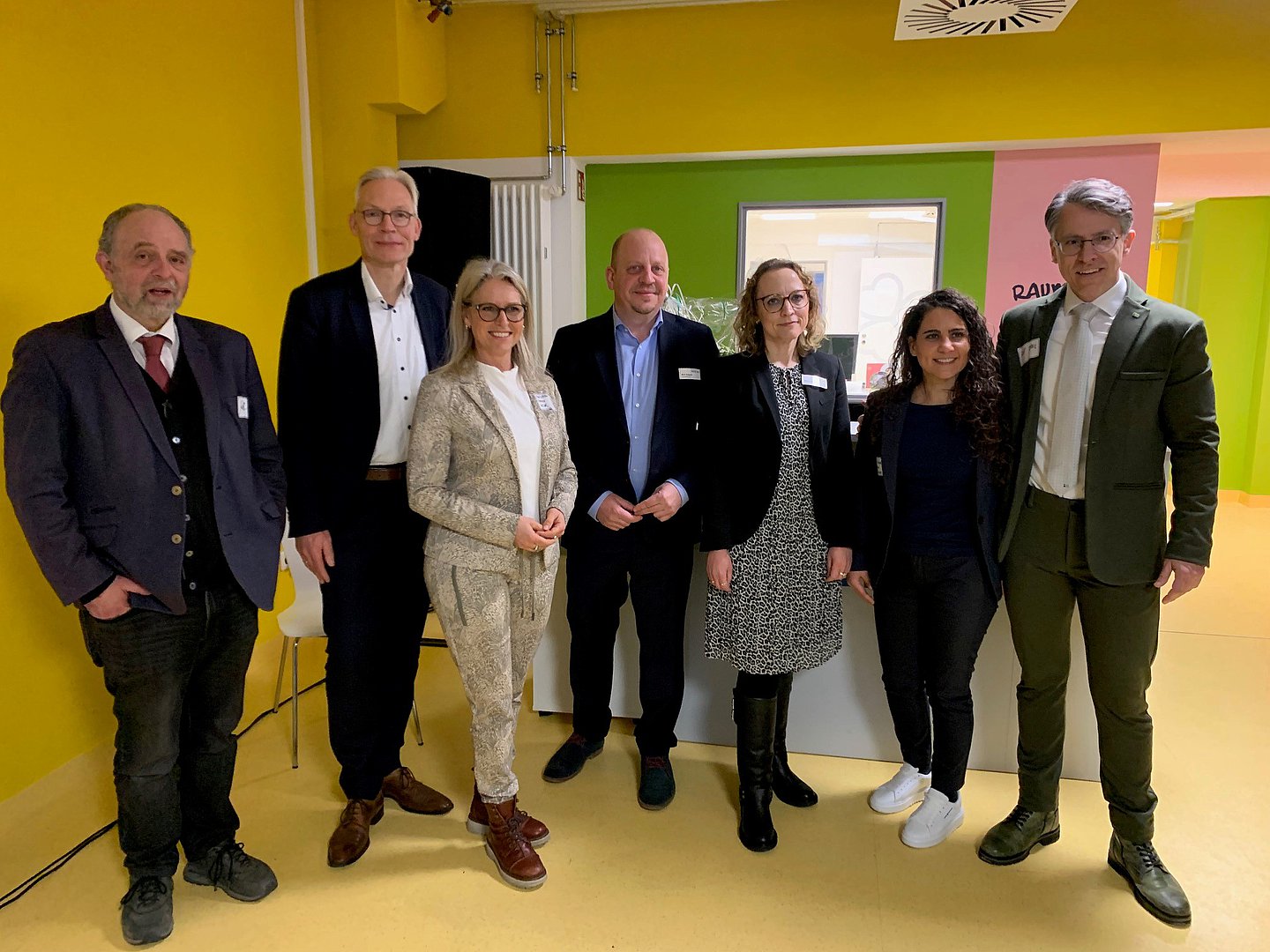 sieben Menschen in Anzügen und Kleidern stehen vor einer Gelben Wand.