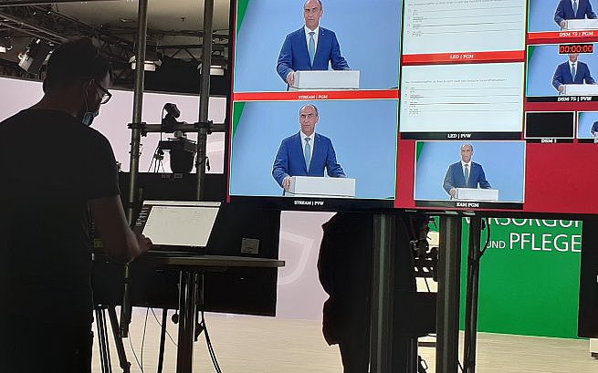Das Bild zeigt die Regie eines TV-Studios. Vor einem großen Flachbildschirm mit zwei großen und sieben kleinen göffneten Computer-Fenstern steht ein Mann mit Brille und Bart an einem Laptop und tippt. Im Hintergrund kann mann das TV-Studio erahnen. 