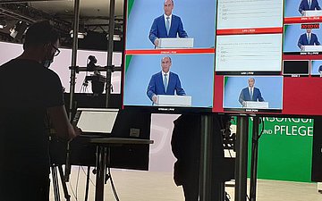 Das Bild zeigt die Regie eines TV-Studios. Vor einem großen Flachbildschirm mit zwei großen und sieben kleinen göffneten Computer-Fenstern steht ein Mann mit Brille und Bart an einem Laptop und tippt. Im Hintergrund kann mann das TV-Studio erahnen.