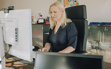 Diana Stolz, Gesundheitsministerin von Hessen, an ihrem Schreibtisch. Sie sitzt auf einem Leberstuhl und schaut auf einen gro&szlig;en Bildschirm. Im Hintergrund ein Bild an der Wand und ein kleiner Schrank, auf dem eine Hessen-M&uuml;tze zu sehen ist.