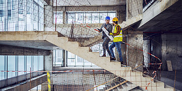 Zwei Bauarbeiter mit Schutzhelmen stehen auf einer Beton-Treppe in einem Rohbau und besprechen Baupl&auml;ne. Um sie herum sind Betonw&auml;nde, gro&szlig;e Fenster und provisorische Absperrungen mit Bewehrungsstahl in einem unfertigen Geb&auml;ude.