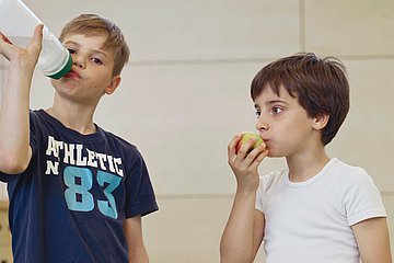 Kindern sollte man früh vermitteln, wie wichtig ausreichend Flüssigkeit für den Körper ist. Mehr zu trinken, kann man mit Trinkritualen wie beispielsweise gemeinsames Trinken beim Essen erlernen.  Foto von zwei Kindern die trinken und Obst essen