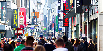 Foto: Menschenmenge in Fu&szlig;g&auml;ngerzone mit bunten Logoschildern der Gesch&auml;fte.