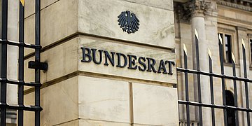 Bundesrat Das Foto zeigt den Eingangsbreich des Bundesrates in Berlin in Nahaufnahme. Zu sehen ist auf einer Säule der Schriftzug BUndesrat in Metalllettern, darüber der Bundesadler. Rechts und links säumt ein Metallzaun mit goldfabrenen Spitzen die Säule.