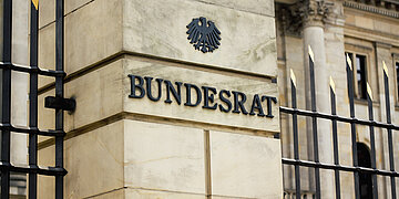 Das Foto zeigt den Eingangsbreich des Bundesrates in Berlin in Nahaufnahme. Zu sehen ist auf einer S&auml;ule der Schriftzug BUndesrat in Metalllettern, dar&uuml;ber der Bundesadler. Rechts und links s&auml;umt ein Metallzaun mit goldfabrenen Spitzen die S&auml;ule.