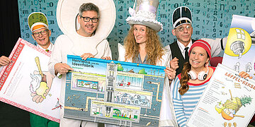 Foto: Das Ensemble des AOK-Kindertheaters "Henrietta" steht auf der B&uuml;hne und h&auml;lt Plakate hoch.
