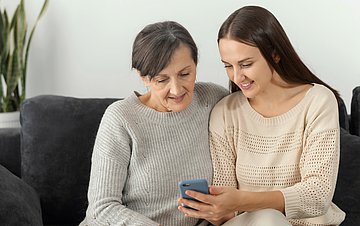 ePA-Infos zur Pflege Foto einer jüngeren Frau, die mit einer alten Dame auf dem Sofa sitzend ins Handy blickt