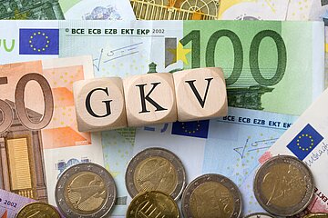 Geldscheine und M&uuml;nzen liegen auf einem Tisch. Auf W&uuml;rfeln stehen die Buchstaben G, K und V.