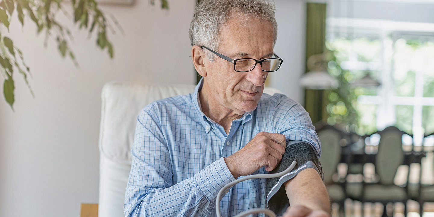 Das Foto zeigt einen Senior im blau-karierten Hemd, der sich am linken Arm die Manschette des Blutdruckmessgerätes anlegt. 