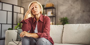 Schwindel Eine Frau mittleren Alters sitzt auf der Couch mit geschlossenen Augen und fasst sich an den Kopf.
