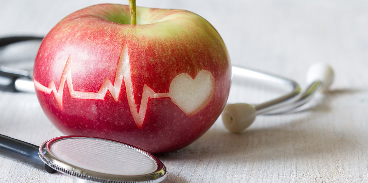 Foto: Neben einem Apfel, auf dem ein Herz-/Kreislaufdiagramm angedeutet ist, liegt ein Stethoskop.