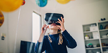 Kind mit einer Virtual-Reality-Brille steht in einem hellen Raum und h&auml;lt die Brille mit beiden H&auml;nden fest; im Hintergrund schweben bunte Luftballons.
