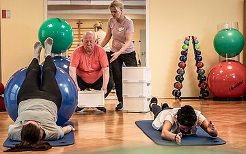 In einem Sportraum machen verschiedene Personen R&uuml;cken&uuml;bungen. Der Raum ist ausgestattet mit B&auml;llen und verschiedenen Sportger&auml;ten. Zwei Personen liegen auf Matten auf dem Boden. Die eine liegt auf dem Bauch und hebt Arme und Beine. Die andere hat die F&uuml;&szlig;e auf einem Gymnastikball und h&auml;lt den K&ouml;rper gerade. Im Hintergrund hilft eine Physiotherapeutin einem Herren bei einer &Uuml;bung.