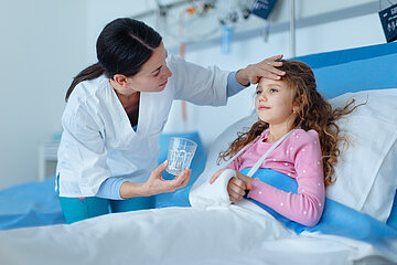 Ein M&auml;dchen liegt mit einem Gipsverband am rechten Arm in einem Krankenhausbett. Eine Krankenschwester hat sich ihr zugewandt und h&auml;lt ihre linke Hand an der Stirn des M&auml;dchens, in der rechten hat sie ein Glas Wasser, das sie dem Kind anbietet.