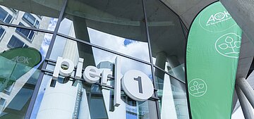 Das Bild zeigt in Nahaufnahme die Glasfront des Eingagsbereichs des Pier 1. Darauf ist die Leuchtaufschrift "Pier 1" und rechts daneben eine gr&uuml;ne Beachflg mit AOK-Logo
