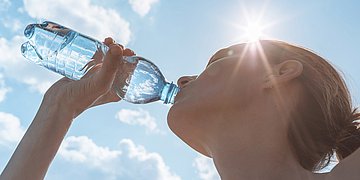 Hitze und Umwelt 1 Das Foto zeigt den in den Ncken gelegten Kopf einer dunkeblonden Frau mit Zopf, die vor sonnigem weiß-blauen Himmel aus einer PET-Flache Wasser trinkt .