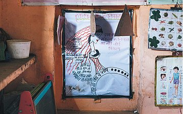 An einer orange gestrichenen Wand hängen verschiedene Poster. Rechts hängen zwei kleinere Poster mit Gemüsesorten und Kinder-Körperteilen drauf. In der Mitte hängt eine große Zeichnung von einer Mutter mit ihrem Baby im Arm. Drum herum indische Schriftzeichen.