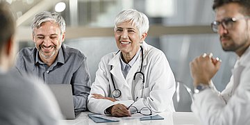 Foto: Mehrere Ärzte sitzen zusammen an einem Tisch und diskutieren bzw. lächeln.