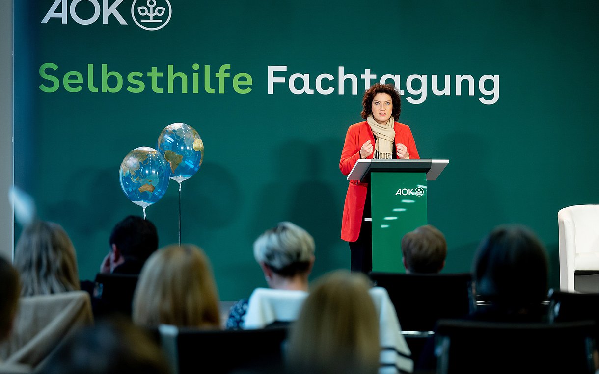 Carola Reimann steht auf einer Bühne vor einem Pult mit Mikrofon. Im Hintergrund steht an der Wand: Selbsthilfe-Fachtagung