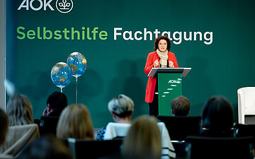 Carola Reimann steht auf einer B&uuml;hne vor einem Pult mit Mikrofon. Im Hintergrund steht an der Wand: Selbsthilfe-Fachtagung