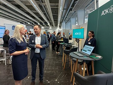 Das Bild zeigt eine blonde Frau in einem blauen Kleid (links) im Gespr&auml;ch mit einem graumelierten Mann mit hoher Stirn in einer Art Messehalle. Rechts sieht man einen LED-Bildschirm mit dem AOK-Logo sowie mehrere Stehtische und Hocker aufgereiht.
