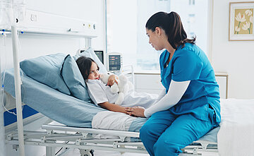 Ein M&auml;dchen liegt mit einem Kuscheltier im Arm in einem Krankenhausbett. Eine weibliche Krankenhausmitarbeiterin sitzt an ihrem Bett und h&auml;lt ihr die Hand.
