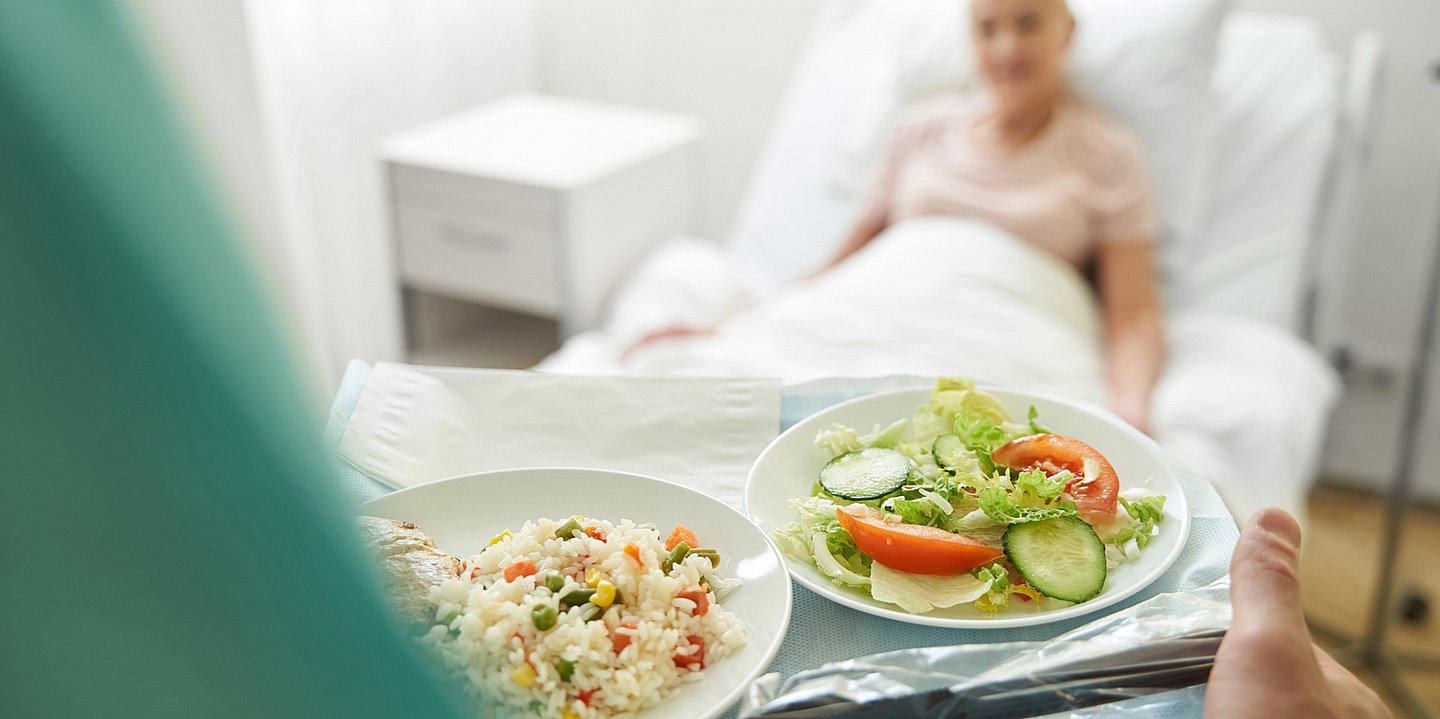 Foto: Eine Pflegeperson trägt ein Tablett mit Essen in der Hand, dahinter liegt eine Patientin im Bett eines Krankenhauses.