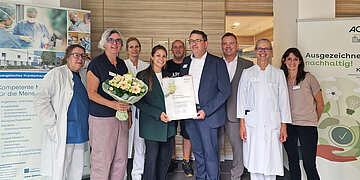 Eine Gruppe aus Menschen, darunter &Auml;rzt*innen, steht vor einem Krankenhaus. Vorn steht Christoph Patzelt, Gesch&auml;ftsf&uuml;hrer des Evangelischen Krankenhauses, der eine Urkunde h&auml;lt, auf der "Krankenhaus Nachhaltigkeitspreis 2026" steht.