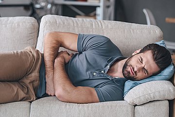 Magen-Darm-Infektionen Ein auf dem Sofa liegender Mann hält sich mit schmerzverzerrtem Gesicht den Bauch.