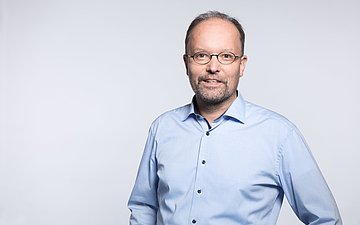 Ralf Metzger Porträtfoto zeigt Kopf und Rumpf, Ralf Metger trägt eine ovale Brille und ein hellblaues Hemd.