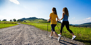 Zwei Frauen beim Gehen mit Walkingst&ouml;cken auf einem sonnigen Feldweg. Die Sonne scheint von einem klaren Himmel, und im Hintergrund sind Wiesen, B&auml;ume und sanfte H&uuml;gel zu sehen.