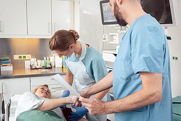 Darmkrebs in Westfalen-Lippe Eine weibliche Patientin liegt auf der Liege eines Behandlungsraums für eine Darmspiegelung. Eine Assistentin legt ihr eine Blutdruckmanschette um den linken Oberarm an und der behandelnde Arzt begrüßt die Patientin per Handschlag. An der Hand sieht man den bereits gelegten Zugang.
