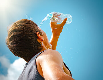 Vor blauem Himmel steht ein junger Mann im armfreien Shirt im Freien und h&auml;lt eine Wasserflasche hoch und vors Gesicht, so dass dieses mit dem herauslaufenden Wasser bespritzt wird.