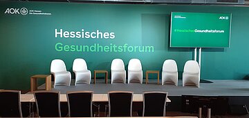 Das Foto zeigt ein leeres Podium mit sechs weis&szlig;en, geschwungenen St&uuml;hlen und drei h&ouml;zernenn Beistelltischchen dazwischen. Im Vordergrund sieht man f&uuml;nf schwarze mit Holz umrandete Stuhllehnen , im Hintergrund den gr&uuml;n-wei&szlig;en Schriftzug &bdquo;Hessisches Gesundheitsforum" auf dunkelgr&uuml;nen Grund. Rechts steht ein gro&szlig;er Flachbildschirm mit dem Schriftzug &bdquo;#HessischesGesundheitsforrum&ldquo; in der gleichen Optik.