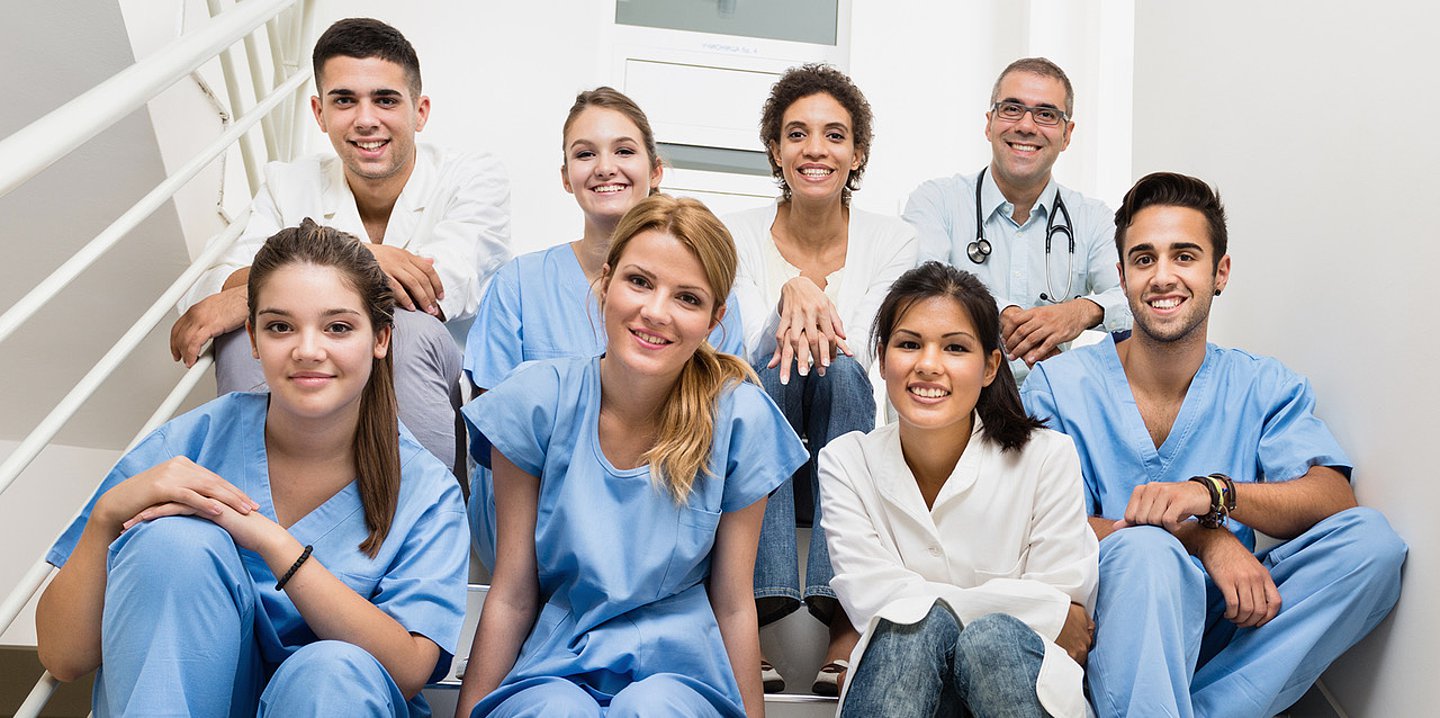 Foto: Eine Gruppe junger Menschen in Pflegekleidung sitzt auf einer Treppe und lächelt.