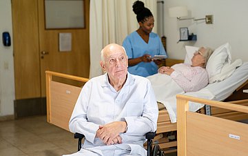 Demenzsensible Behandlung Krankenhauszimmer mit einem älteren Mann auf einem Stuhl im Vordergrund. Im Hintergrund spricht eine Pflegerin mit einer alten Dame im Bett.